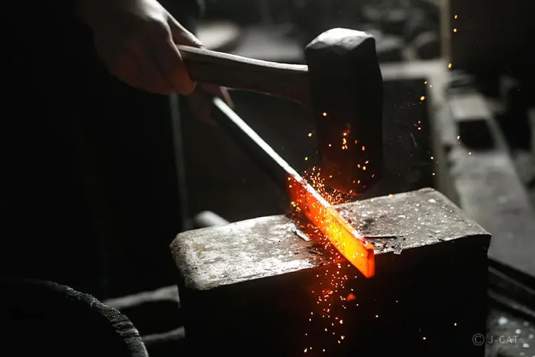 experiences_gifu_knife-making-workshop_hammering-red-hot-steel A close-up, action shot of a participant's hands rhythmically striking a glowing, red-hot tamahagane steel billet against an iron anvil with a heavy hammer, sending bright orange sparks flying during the high-heat hammer forging step.