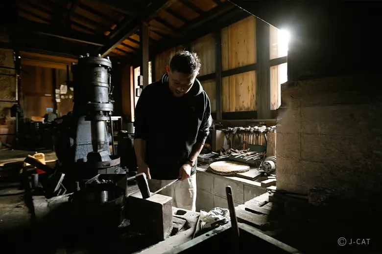 experiences_gifu_knife-making-workshop_swordsmith-hammering-steel-in-forge-with-tools Wide shot of the authentic Asano Kajiya swordsmith forge, showing the master smith standing by the anvil with heavy machinery, iron hammers, and specialized bladesmithing tools arranged in the dim, atmospheric workshop.