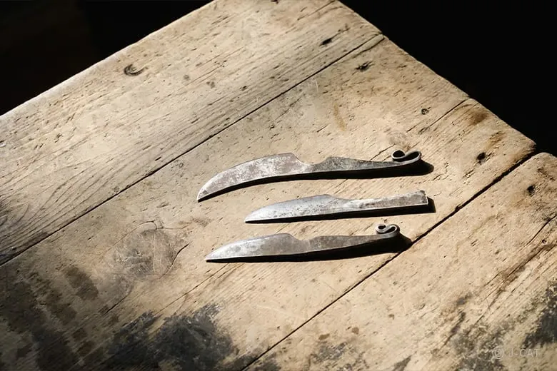 experiences_gifu_knife-making-workshop_three-example-designs-of-newly-forged-blades Three unpolished, raw examples of newly forged utility knife blades resting on a light wooden surface, showcasing different shapes, classic profiles, and decorative curled tang designs before final hilt finishing.