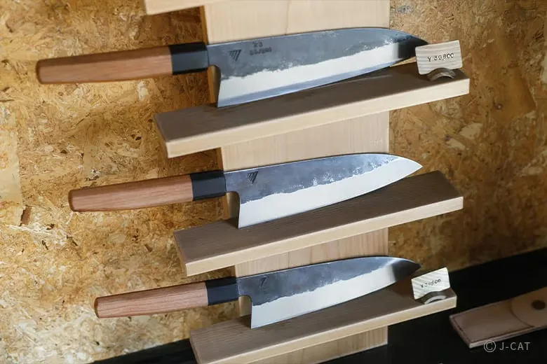 experiences_gifu_knife-making-workshop_three-finished-knives-with-wooden-handles Three completed, custom-forged knives displayed on a wooden stand, each finished with a sleek, minimalist wooden handle, a dark bolster, and a visible wavy blade pattern (hamon) resulting from clay coating and water quenching.