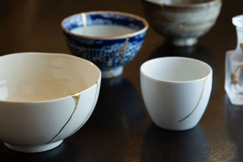 A curated collection of genuine, premium Japanese ceramics and glass tableware neatly displayed on a dark surface, each showcasing beautiful golden lacquer seams from kintsugi repairs that highlight the wabi-sabi philosophy of celebrating imperfections.