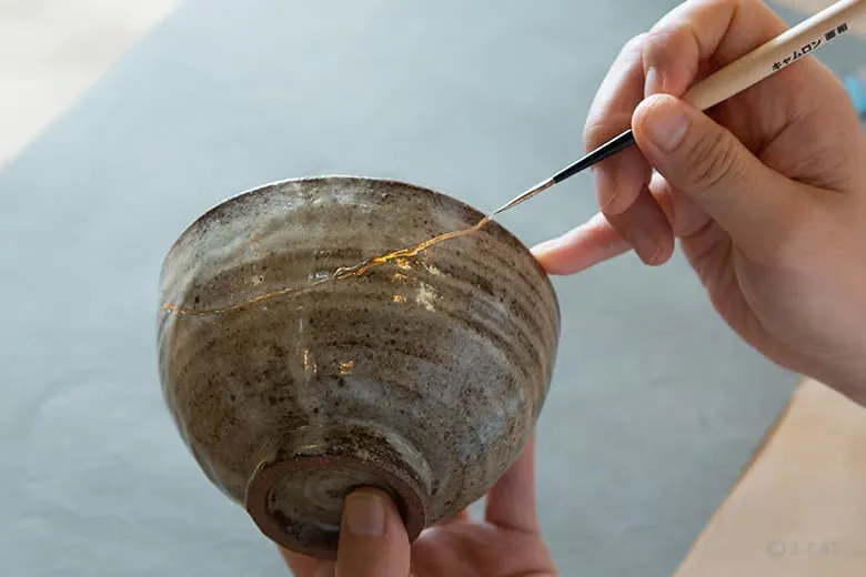 A close-up view of a participant’s hands during the workshop, delicately using a fine-tipped artisan paintbrush to trace a fracture line with golden lacquer across the surface of an earth-toned Japanese ceramic bowl.