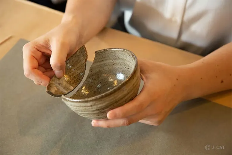 Close-up shot of a participant’s hands carefully fitting two pieces of a broken ceramic bowl back together over a gray work mat, demonstrating the initial step of treating fractures and securely joining the components.