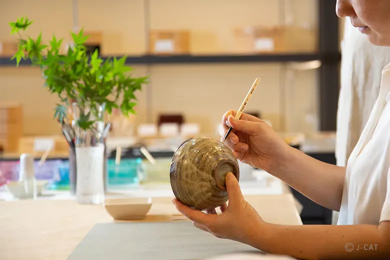 Side profile of a Japanese woman holding a rustic ceramic bowl upside down, using a fine craft brush to apply gold powder along the repaired crack lines in a bright, organized workshop space with green foliage in the background.