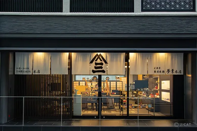 The illuminated storefront of the century-old Utsuwa Nihonbashi Mutoh craft house at night, featuring clean glass entry doors, a modern guardrail, and traditional white fabric noren curtains printed with bold black Japanese characters.