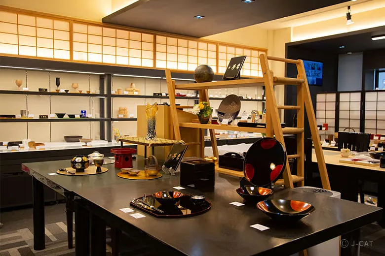 The elegant interior showroom of Utsuwa Nihonbashi Mutoh, showing wide wooden display tables and well-lit background shelves showcasing high-quality Japanese lacquerware, Arita-yaki, and Kutani-yaki ceramic collections under traditional warm lighting.