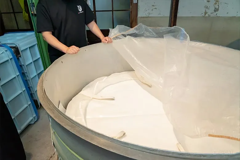 An indoor shot at Sasaki Shuzo showing a brewer standing next to a massive open-topped fermentation vat lined with protective sheeting, showcasing the active production facility floor where yeast transforms starches into premium Kyoto sake.
