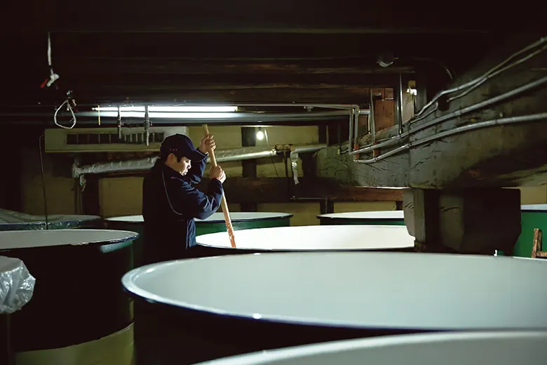 Inside the historic earthen-walled cellars of Sasaki Shuzo, a kurabito (brewer) clad in a cap uses a long wooden oar to methodically stir a large, active vat of bubbling white moromi sake mash beneath traditional timber beams.