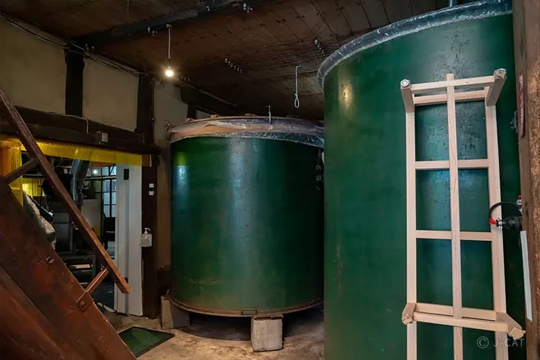 The atmospheric inner production floor of Kyoto’s last central independent brewery, Sasaki Shuzo, displaying large green stainless steel conditioning tanks and a wooden ladder under a dim light bulb to preserve ancestral Rakuchu brewing traditions.