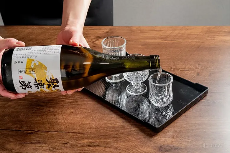 Close-up of a host carefully pouring crisp, clear Jurakudai premium sake from a dark bottle into fine, cut-glass tasting cups on a black lacquer serving tray during the private tasting experience at Sasaki Shuzo in Kyoto.