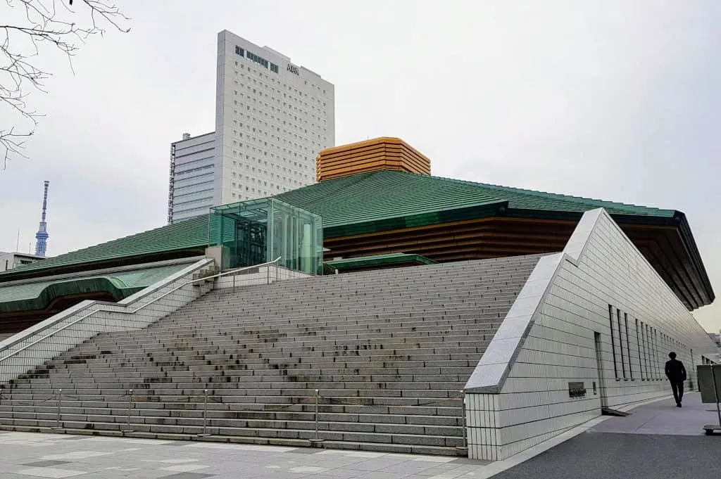 experiences_tokyo_ryogoku-sumo-tournament-arena Wide exterior view of the iconic Ryogoku Kokugikan sumo tournament arena in Tokyo, featuring its large green tiered roof, wide concrete grand staircase, and Tokyo Skytree visible in the distant background.