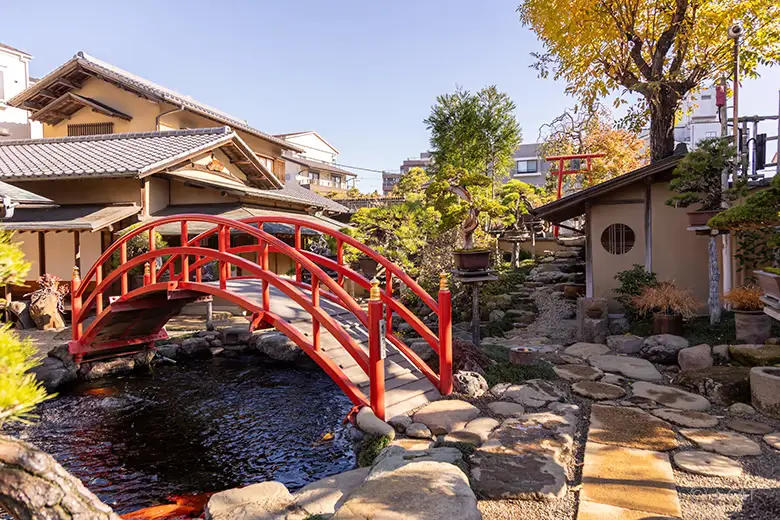 experiences_tokyo_shunkaen-bonsai-worksho-traditional-japanese-garden-drum-bridge-over-koi-pond Wide view of the pristine outdoor gardens at the Shunkaen Bonsai Museum, showcasing an iconic arched red wooden drum bridge built over a stone-lined koi pond, with stone paths winding past tiled-roof traditional buildings.