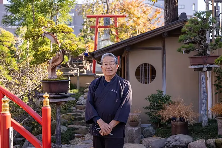 experiences_tokyo_shunkaen-bonsai-workshop-founder-kunio-kobayashi Shunkaen Bonsai Museum founder and legendary master Kunio Kobayashi standing in a traditional navy uniform, holding pruning shears in a sunlit courtyard garden beside an old miniature specimen tree and a red torii gate.