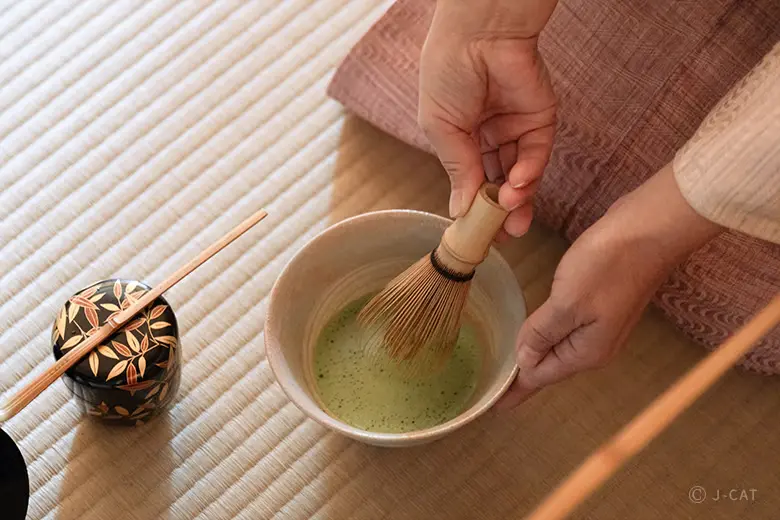 experiences_tokyo_shunkaen-bonsai-workshop-japanese-tea-ceremony Close-up of hands using a traditional bamboo whisk to mix fresh, frothy matcha green tea inside a ceramic bowl on a tatami mat next to a lacquer tea caddy decorated with gold leaf.