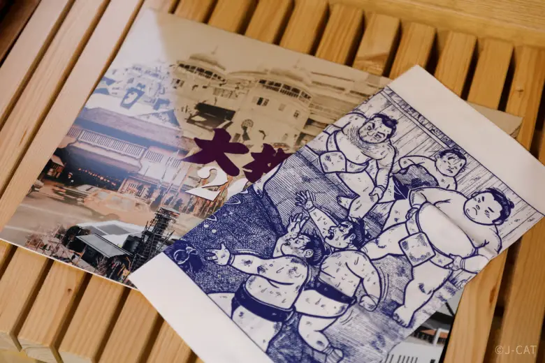 experiences_tokyo_sumo-experience-information-guides Educational sumo information guides and traditional ukiyo-e style woodblock print brochures resting on a light wooden bench, provided during the tour to help guests understand wrestler diets, rules, and Shinto customs.
