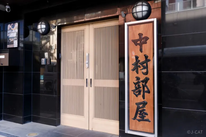 experiences_tokyo_sumo-practice-stable-entrance The discreet, elegant exterior entrance of an active, functioning Tokyo sumo stable (heya), featuring light wood-slatted double doors and a traditional vertical wooden sign displaying bold black calligraphy characters mounted on a polished dark stone wall.