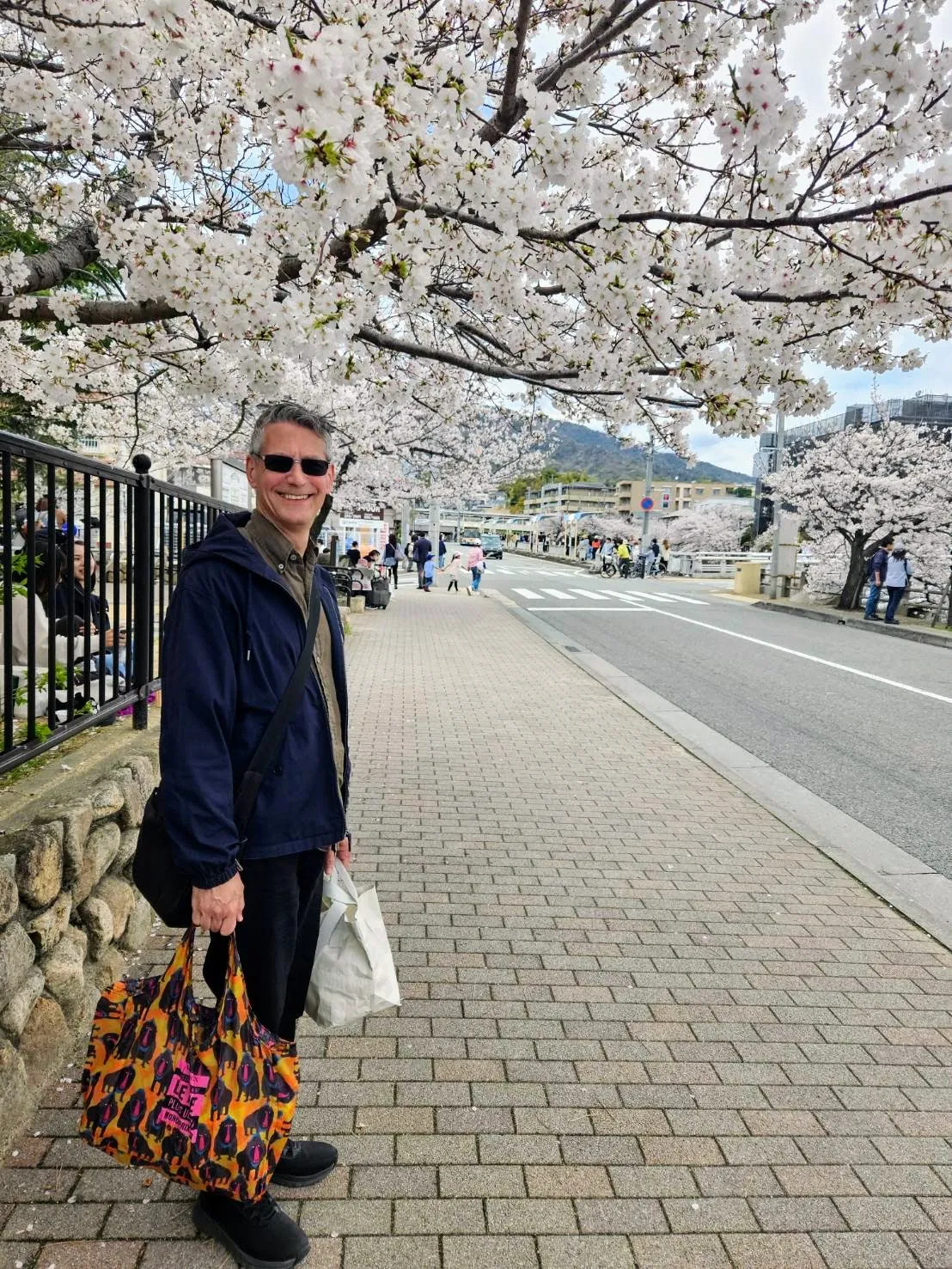 Rob Dyer, founder of the independent travel guide The Real Japan, smiling on a brick sidewalk beneath a canopy of blooming white cherry blossoms (sakura) along a tree-lined street in Ashiya City during spring.