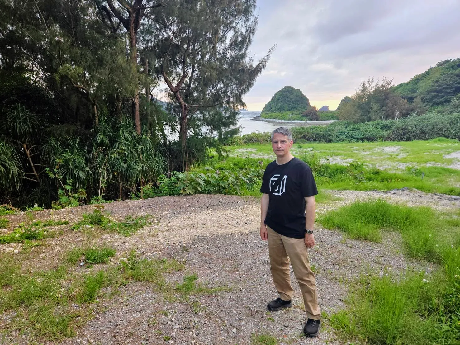 Rob Dyer, travel consultant and founder of The Real Japan, standing in a graphic black t-shirt and khaki trousers on a gravel path overlooking a lush, forested coastline and green sea stacks on Zamami Island.