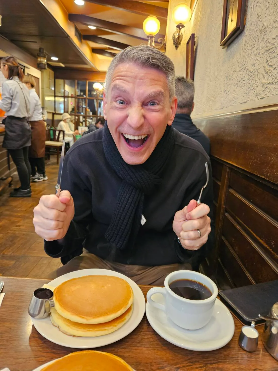 A humorous, expressive portrait of Rob Dyer, founder of The Real Japan, excitedly holding a knife and fork over a classic thick pancakes and hot black coffee inside the historic Santos cafe in Motomachi, Kobe.