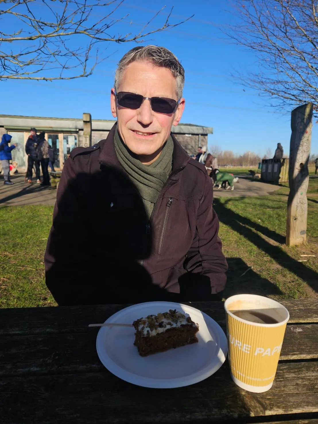 Portrait of Rob Dyer, founder of travel consultancy The Real Japan, wearing sunglasses and a winter jacket at a wooden picnic table with a slice of carrot cake and a takeaway cup of hot coffee at the Jeskyns Community Woodland, Kent UK