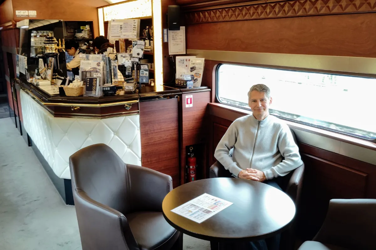 The Real Japan founder Rob Dyer seated inside a premium, retro-styled lounge carriage of the Blue Symphony luxury sightseeing train running between Osaka and Yoshino, featuring dark wood paneling and custom leather armchairs.