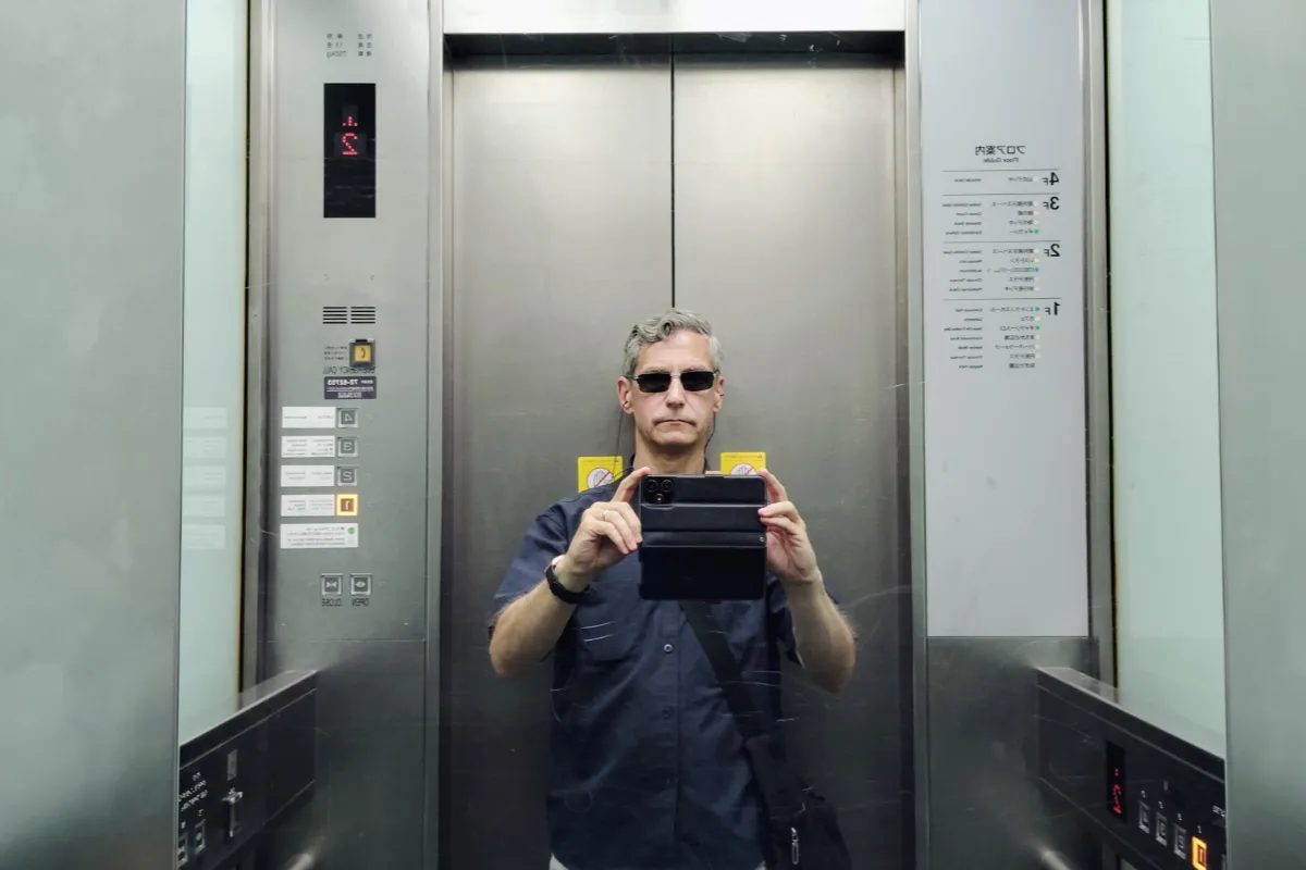 A mirrored elevator selfie of travel specialist Rob Dyer wearing sunglasses and a short-sleeved navy button-down shirt while tracking independent travel research across boutique accommodations in Japan.