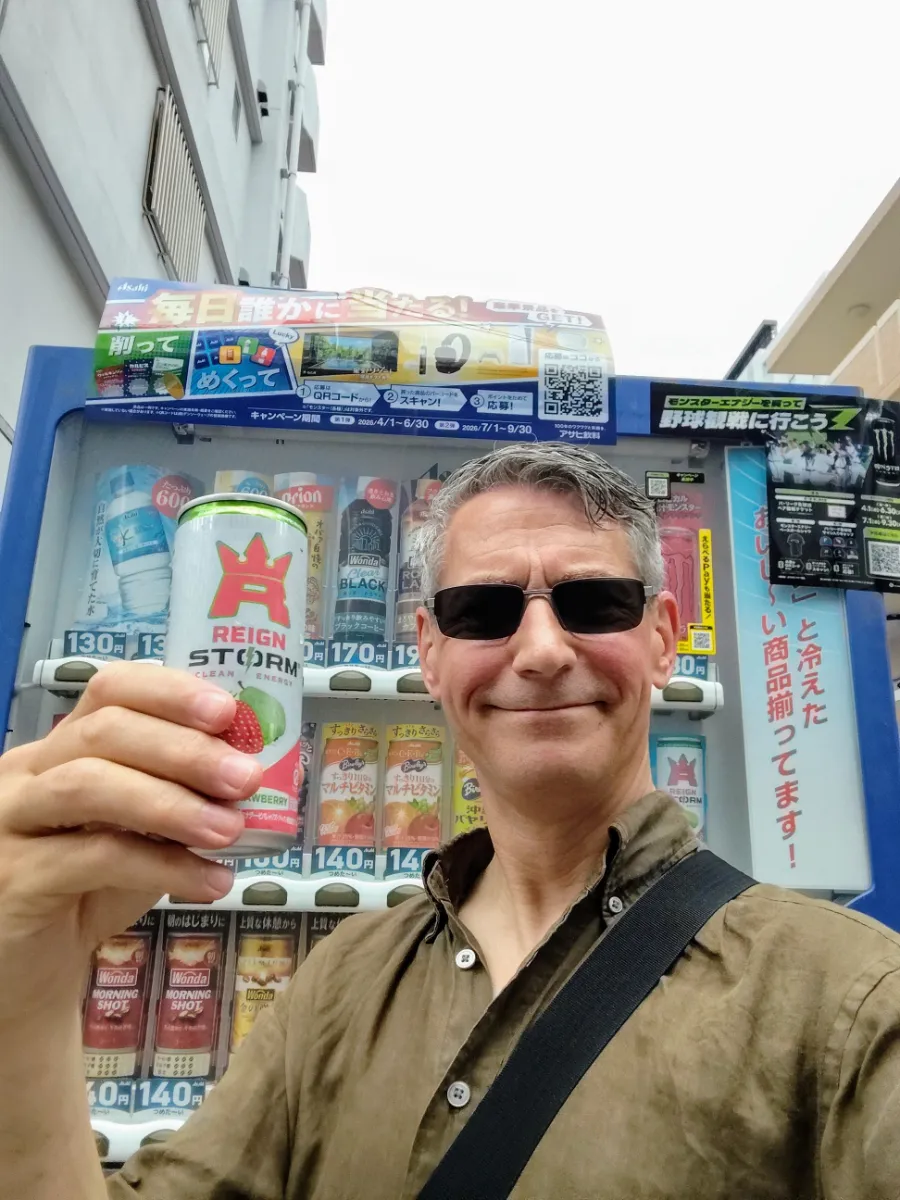 Close-up selfie of Rob Dyer, creator of The Real Japan, smiling while holding up a Reign Storm energy drink in front of a colourful, traditional Japanese beverage vending machine on a city street in Naha Okinawa.