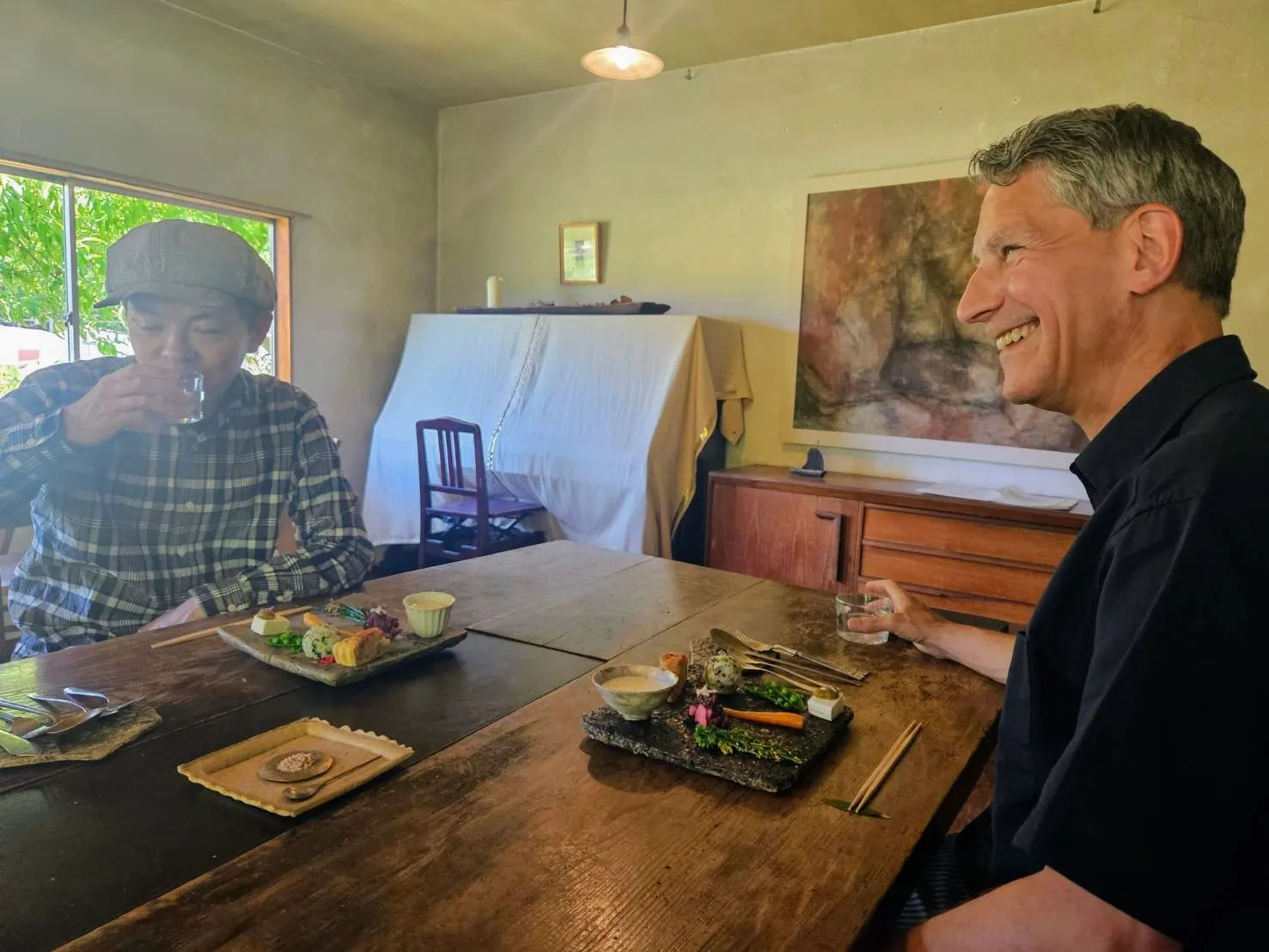 Rob Dyer, founder of The Real Japan, laughing during an off-the-beaten-path culinary experience at a large wooden dining table opposite a friend wearing a flat cap in a minimally designed room at Cafe Millet, Koyto.