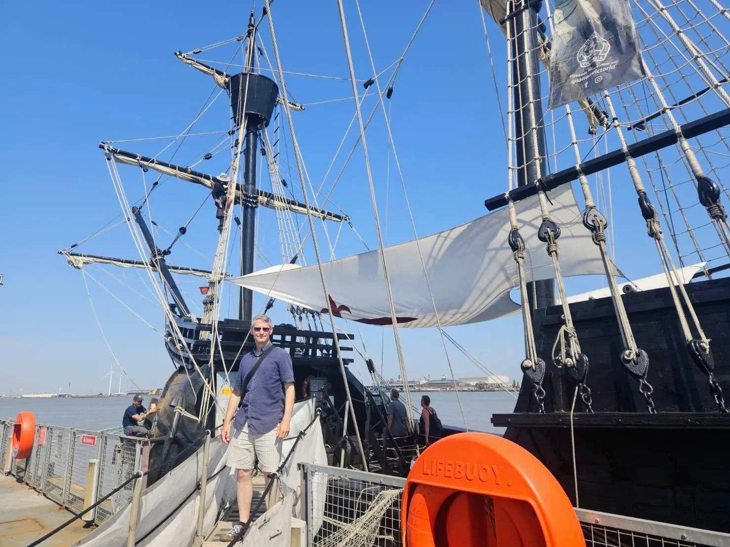 Travel consultant Rob Dyer standing on the gangway of the Nao Victoria a large, rigged historical wooden galleon ship under a clear blue sky, wearing shorts and a short-sleeved shirt during a coastal research trip.