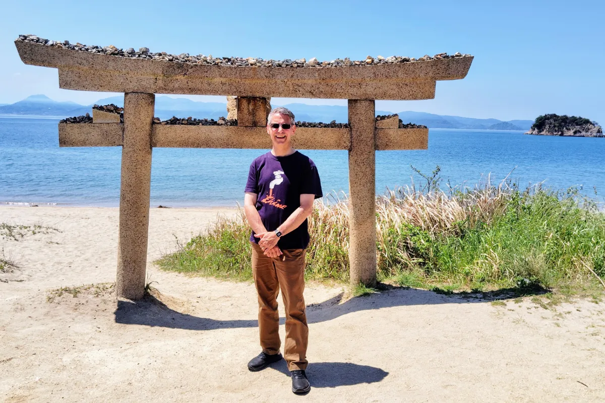 Rob Dyer, independent travel consultant and founder of The Real Japan, smiling on a sandy beach on Naoshima Island under a bright blue sky, standing directly in front of a rustic, stone torii gate covered with small stacked rocks.