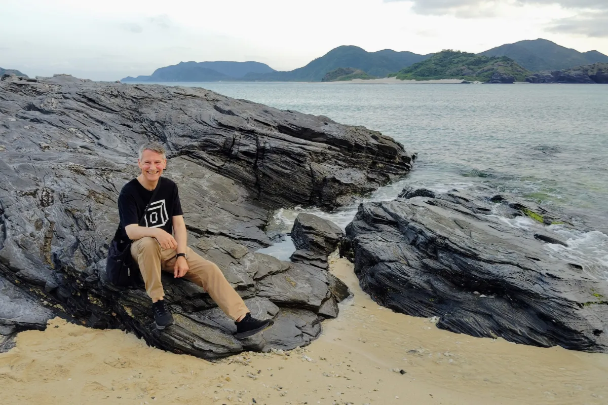 A portrait of Japan travel specialist Rob Dyer sitting and smiling on large, dark volcanic coastal rocks where the tide meets the sand on Zamami Island, Okinawa, with green mountains in the far background.