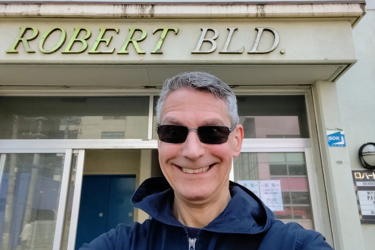 A close-up selfie of Rob Dyer, creator of The Real Japan, wearing sunglasses and a navy blue hooded jacket, smiling in front of the entrance of the "ROBERT BLD." sign in Kobe, Japan.