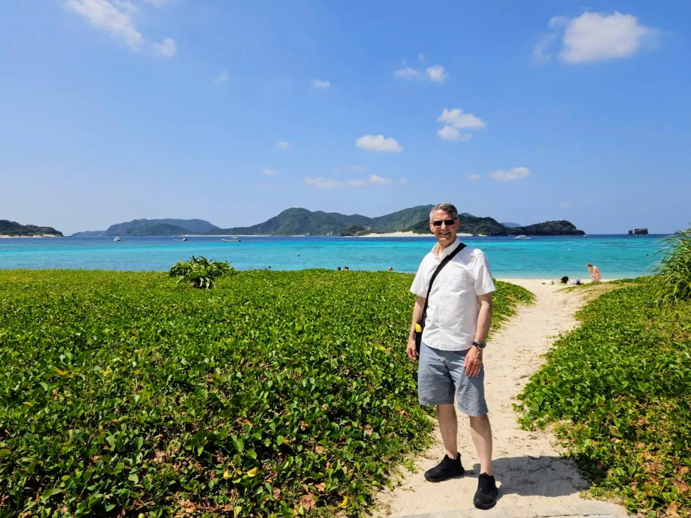 Full-length portrait of Rob Dyer, founder of travel guide The Real Japan, wearing a white short-sleeved button-down shirt and grey shorts on a sandy path surrounded by green foliage, with the brilliant turquoise waters of Zamami Island behind him.