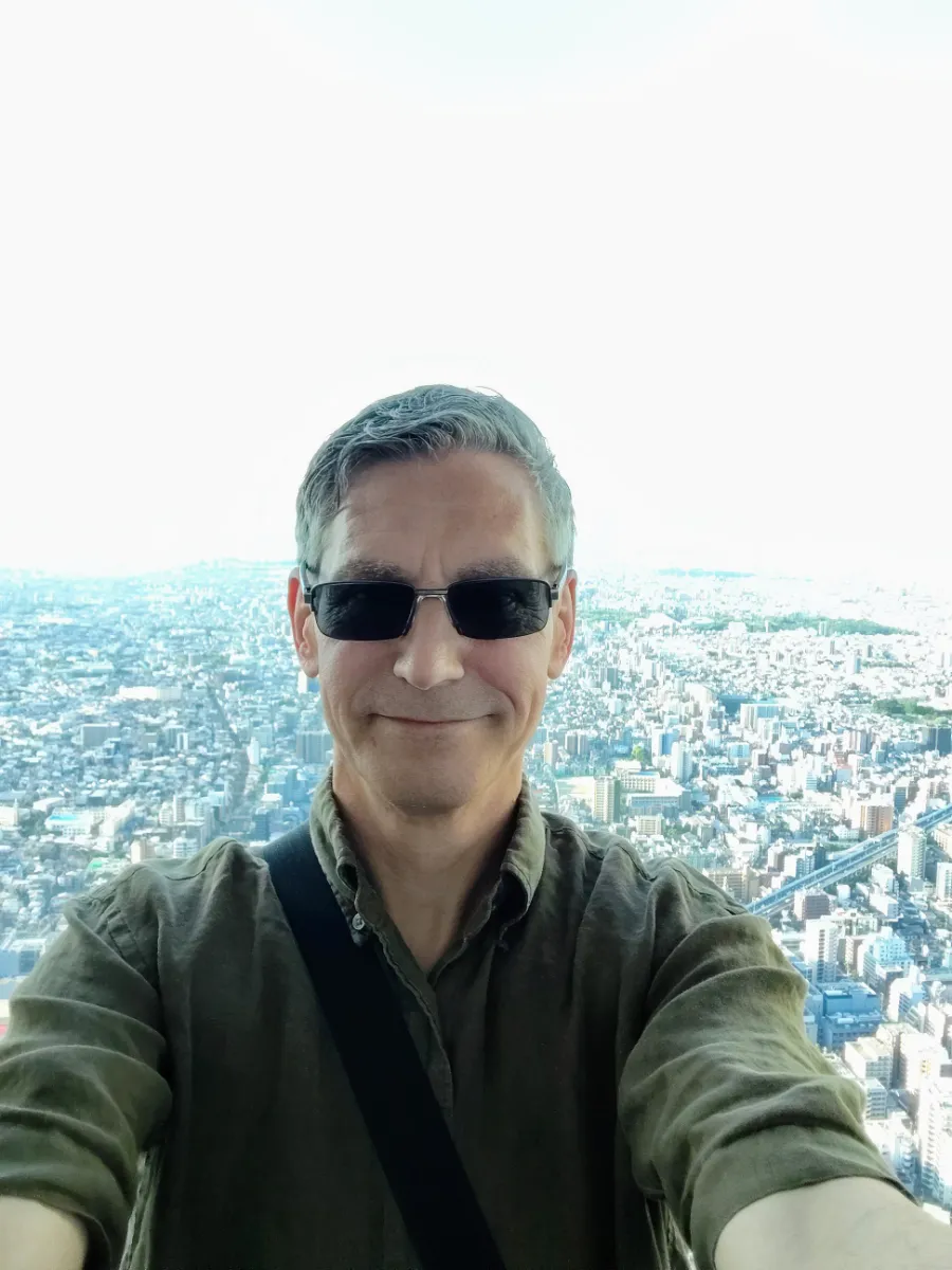 A high-angle selfie of Rob Dyer wearing sunglasses and an olive green shirt, looking out with a calm expression against the expansive, panoramic urban cityscape of Osaka from the high-altitude glass viewing deck at Harukas 300.