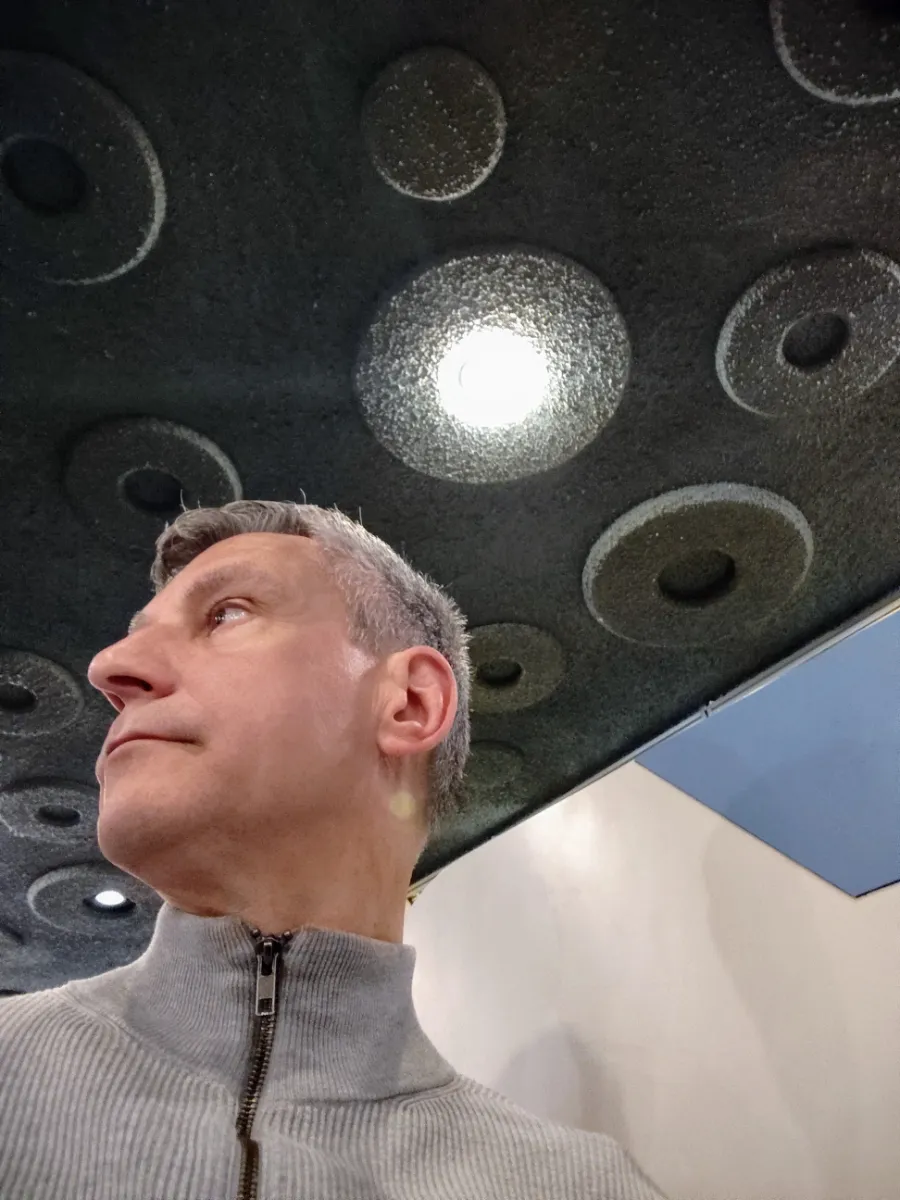 A low-angle portrait looking up at Rob Dyer's profile, founder of The Real Japan, wearing a grey zip-up collar sweater inside the retro-styled Madura cafe in Osaka, highlighting the textured, circular geometric patterns on the dark ceiling.