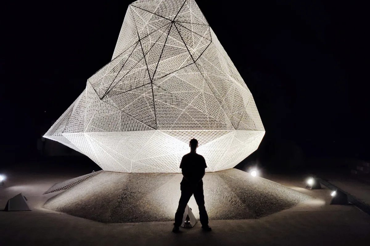 Silhouette of Rob Dyer standing at night in front of the brightly illuminated, white mesh geometric structure of the Naoshima Pavilion, designed by architect Sou Fujimoto on Naoshima Island.