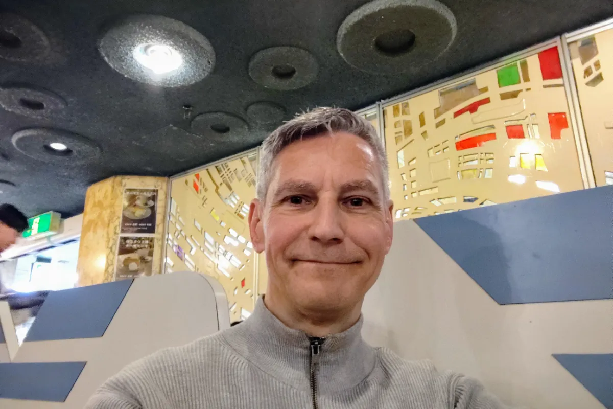 A close-up selfie of a smiling Rob Dyer, travel guide creator for The Real Japan, inside the authentic, mid-century retro Osaka cafe Madura featuring a textured grey ceiling and decorative, multi-colored geometric glass panels in the background.