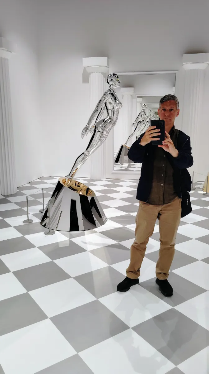 Rob Dyer, founder of The Real Japan, taking a mirror selfie inside a striking museum gallery with a checkered floor, standing next to a sleek, metallic silver retro-futuristic humanoid robot sculpture by Hajime Sorayama at Creative Museum Toyko.