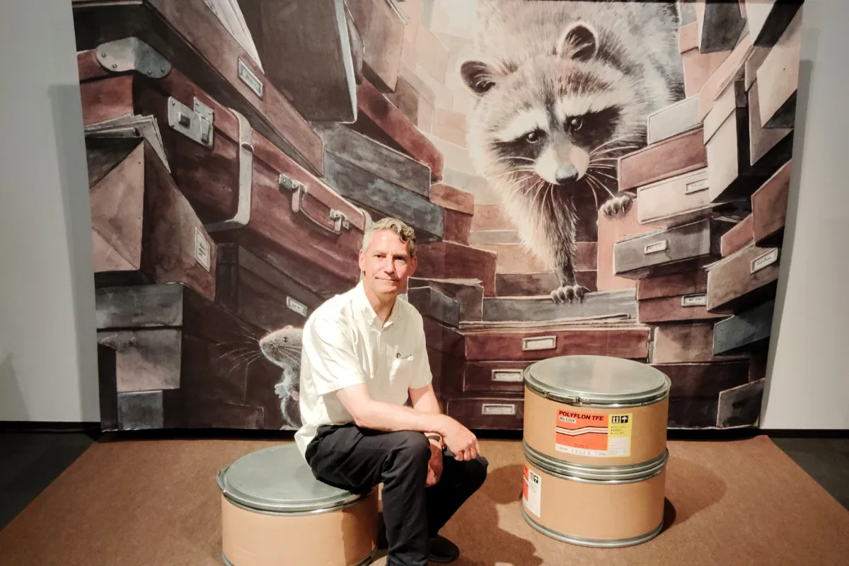 Rob Dyer, founder of independent travel guide The Real Japan, sitting on a cylindrical drum stool in front of a massive, whimsical Torben Kuhlmann illustration backdrop featuring a raccoon and old suitcases at the Kobe Fashion Museum on Rokko Island.