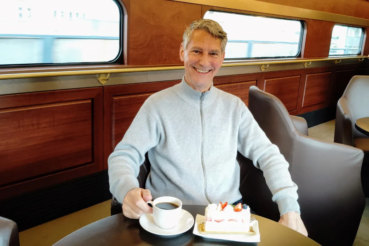 Japan travel specialist Rob Dyer smiling inside the wood-paneled lounge car of the Kintetsu Blue Symphony sightseeing train, holding a white cup of black coffee next to a slice of fresh strawberry shortcake.