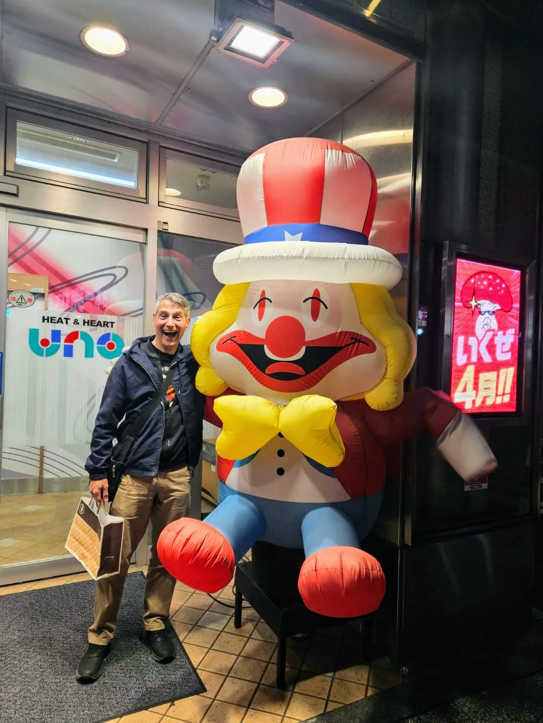 An energetic portrait of Rob Dyer, creator of The Real Japan, laughing and holding a shopping bag next to a large, vibrant, patriotic-themed inflatable clown outside a building at night in Tokyo.