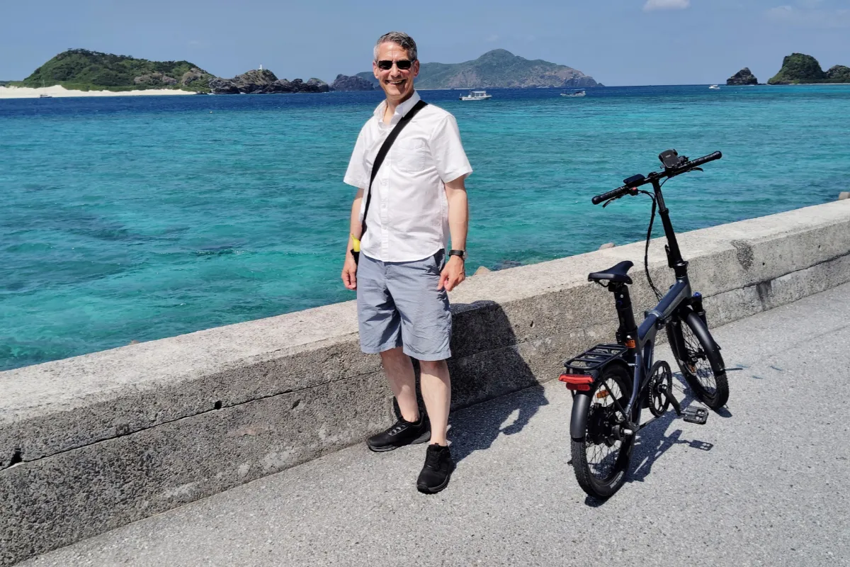 Travel consultant Rob Dyer standing on a paved coastal path next to a dark grey matte folding e-bike on Zamami Island, Okinawa, with brilliant tropical turquoise waters and island hills filling the background.