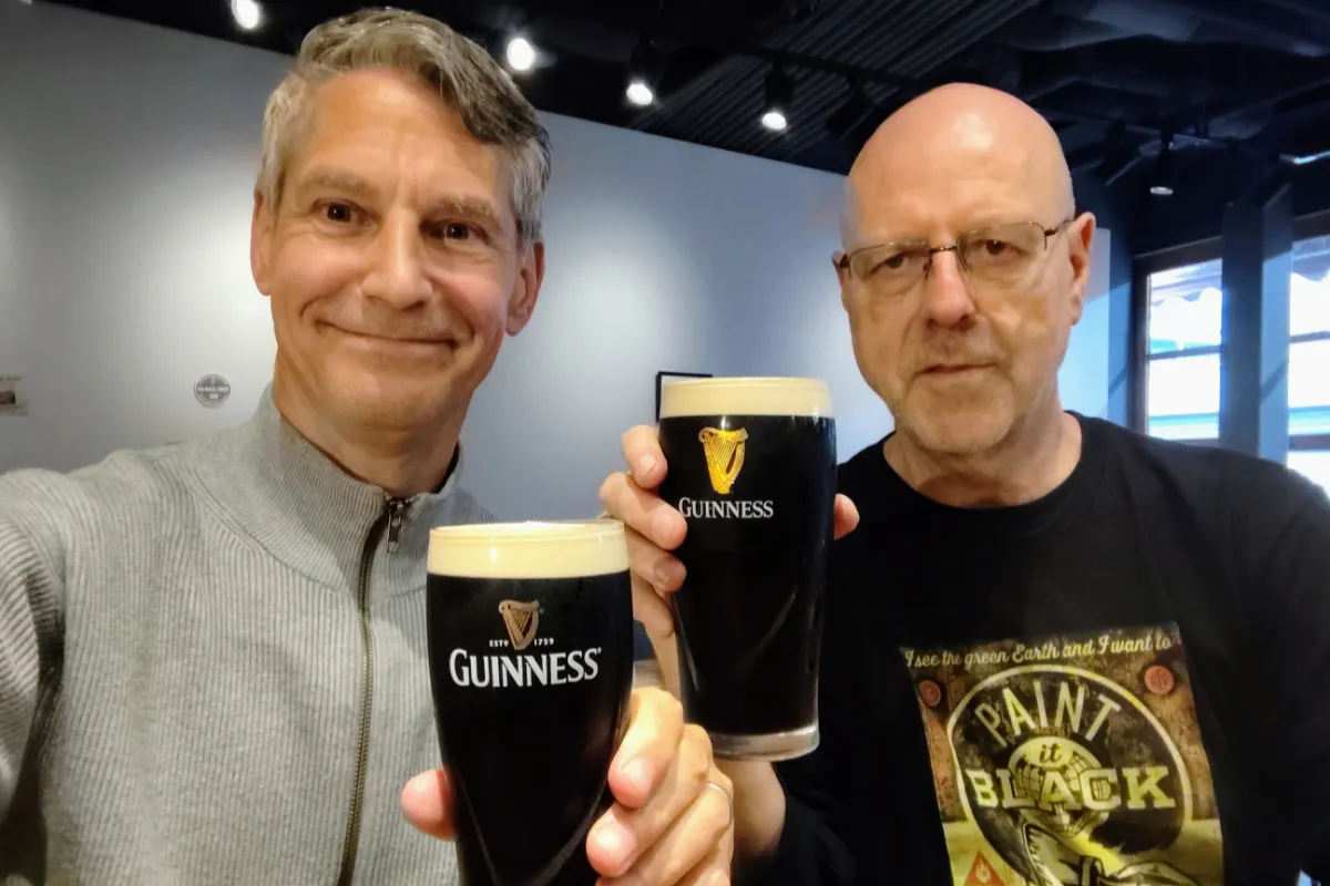 A close-up selfie of Rob Dyer, creator of The Real Japan, smiling as he clinks freshly poured pints of Guinness stout with his friend John Costello inside the British-style Clapham Inn pub in Ashiya, Japan.