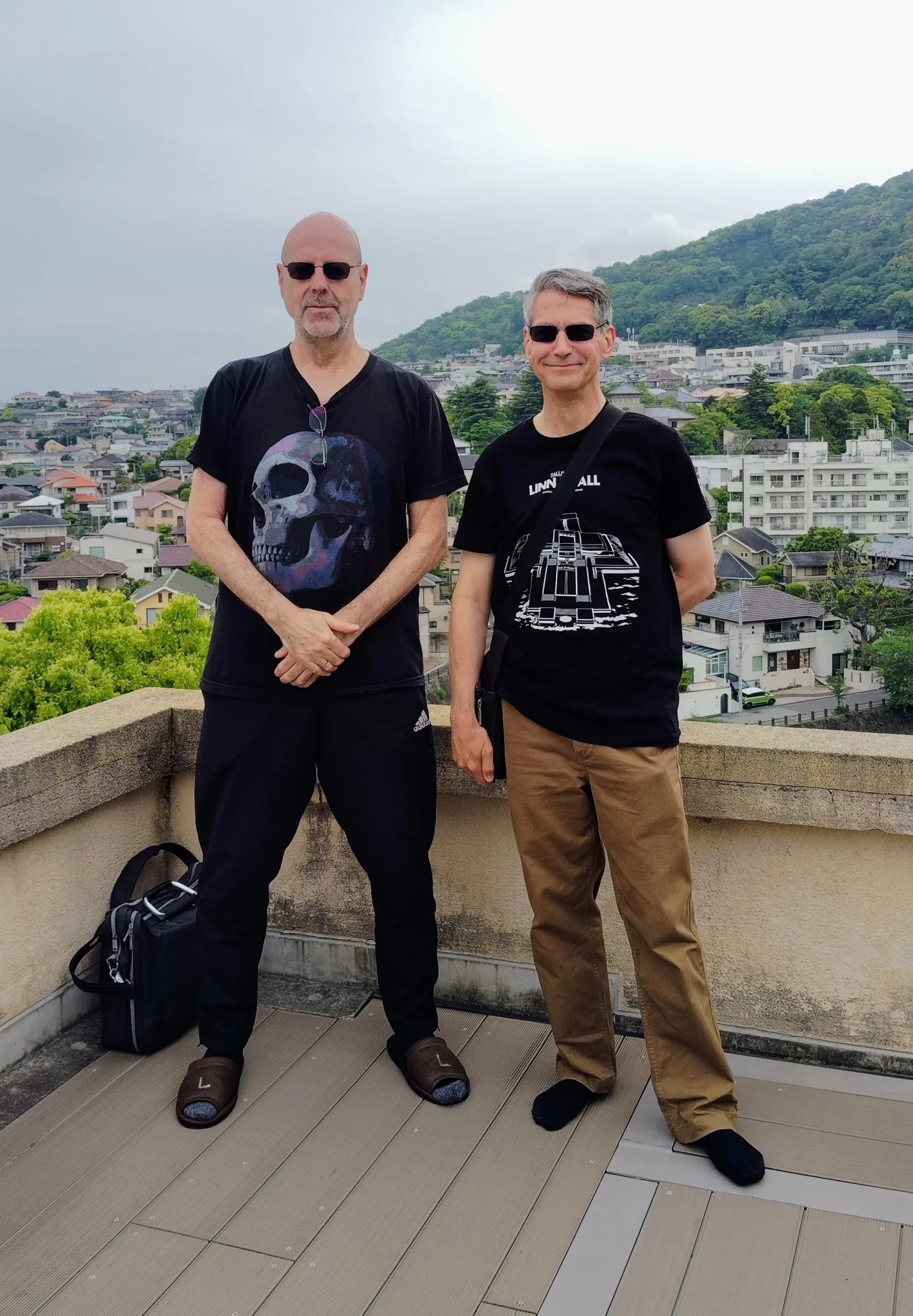 Rob Dyer and his friend John Costello standing on the viewing roof terrace of the historic Yodoko Guest House, an architectural landmark designed by Frank Lloyd Wright in Ashiya, Japan, with the hillside city spreading behind them.