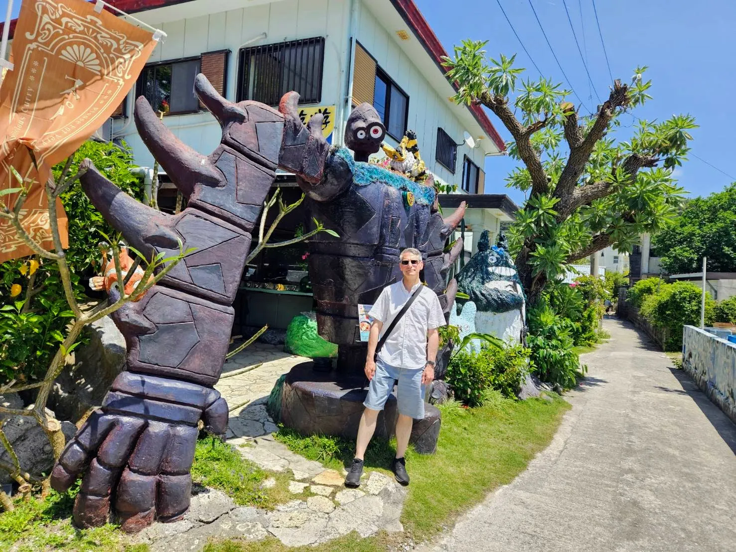 Full-length portrait of Rob Dyer standing next to a large, hand-built sculpture of the iconic Laputa robot from Studio Ghibli's Castle in the Sky, alongside a Totoro figure, outside a local building on Zamami Island.