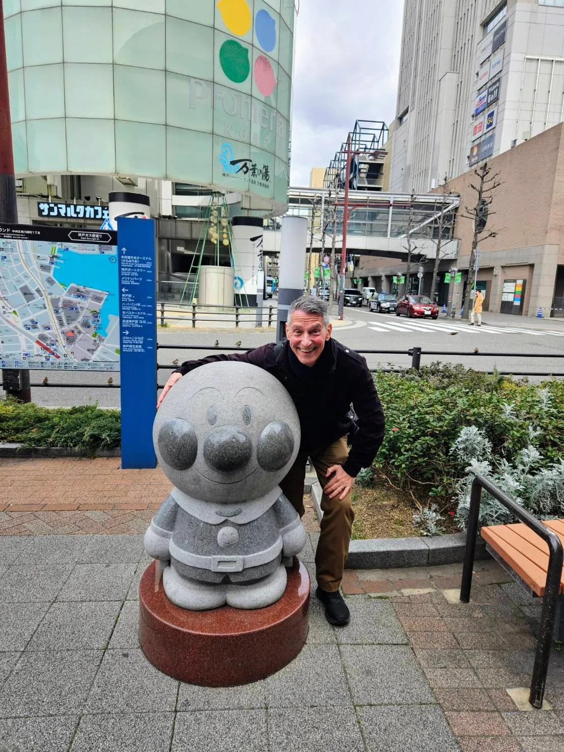 A joyful portrait of travel guide author Rob Dyer leaning over and smiling beside a smooth stone statue of the beloved Japanese anime character Anpanman on a sidewalk in Kobe, Japan.