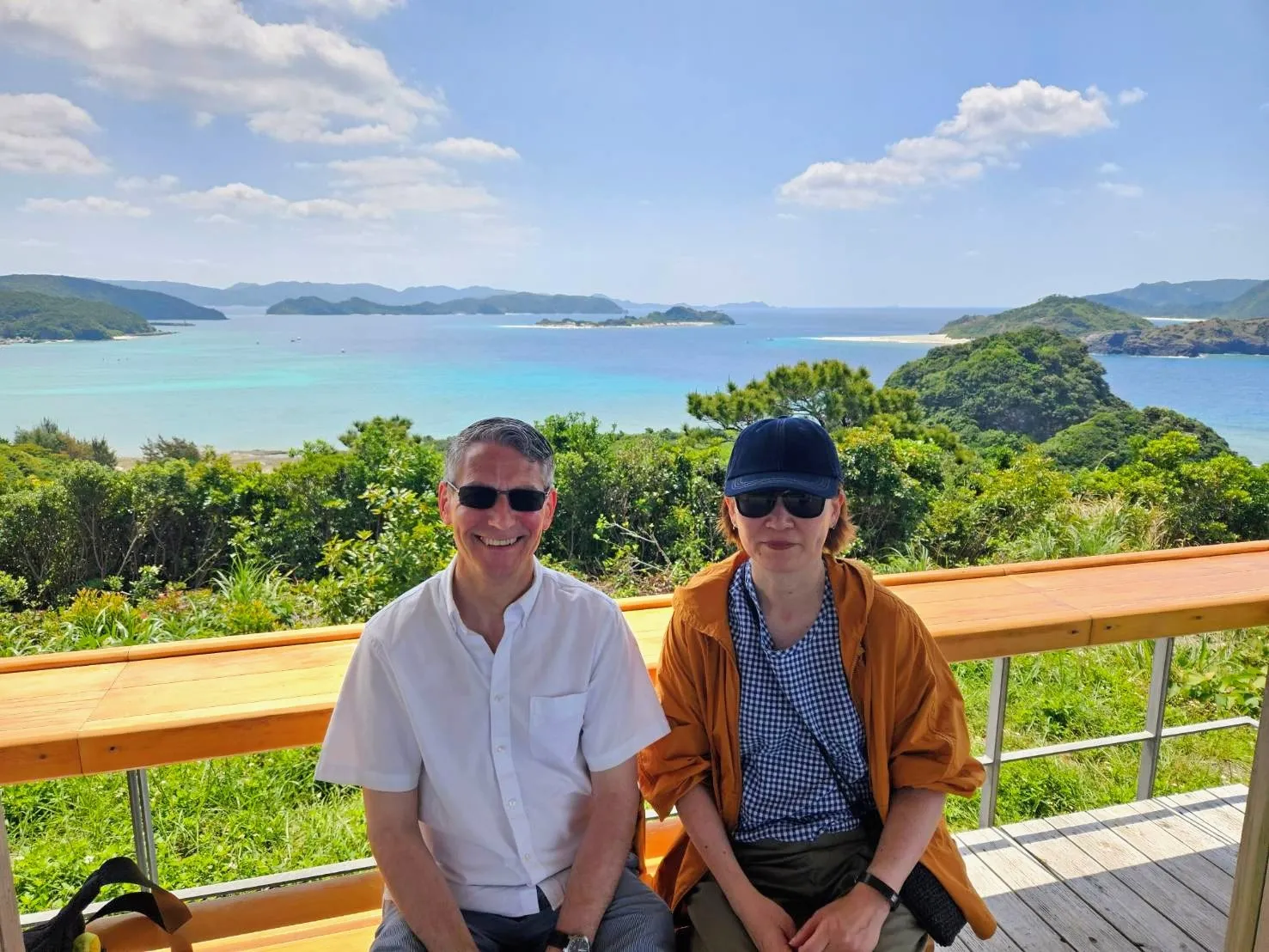 Rob Dyer, independent travel consultant and founder of The Real Japan, sitting smiling with his wife on a wooden observation deck overlooking the breathtaking turquoise waters, tropical coral reefs, and green islands of Zamami, Okinawa, under a bright blue sky with scattered clouds.