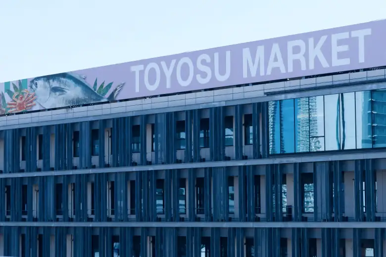 toyosu-fish-market-tour-exterior-with-sign-in-english Exterior wide shot of the modern, state-of-the-art Toyosu Market building facade in Tokyo, featuring a large purple rooftop banner with the name "TOYOSU MARKET" spelled out clearly in English