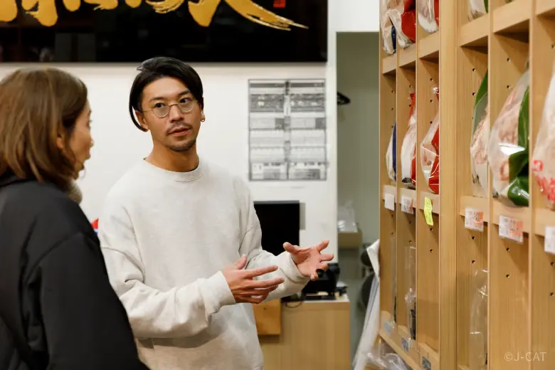 toyosu-fish-market-tour-guide-showing-guest-inside A veteran seafood industry guide wearing glasses gestures with his hands while speaking to a tour guest in front of a wooden display case filled with packaged specialty seafood goods inside a market stall
