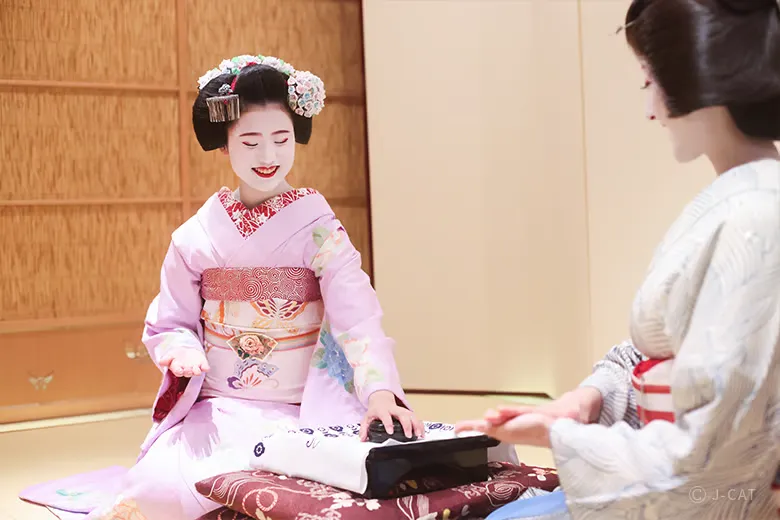 experiences_kyoto_private-geisha-entertainment-dinner-gion-kyoto_maiko-playing-konpira-fune-fune-party-game A smiling maiko and a geiko playing the lively traditional drinking game Konpira Fune Fune over a cushioned box, demonstrating authentic ozashiki asobi parlor entertainment at a private Gion dinner.